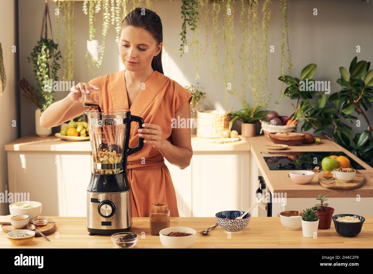 Junge Frau, die beim Kochen in der Küche alle Zutaten für hausgemachten Smoothie oder Dessert in einen elektrischen Mixer gibt Stockfoto