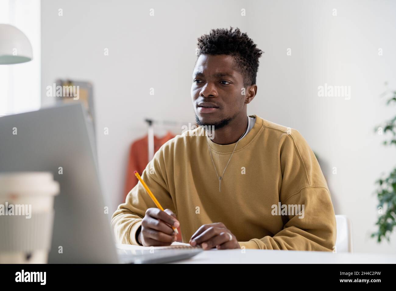 Angespannter junger Mann mit Bleistift, der während des Online-Unterrichts Notizen macht und dem Lehrer zuhört, auf den Laptop-Bildschirm schaut Stockfoto
