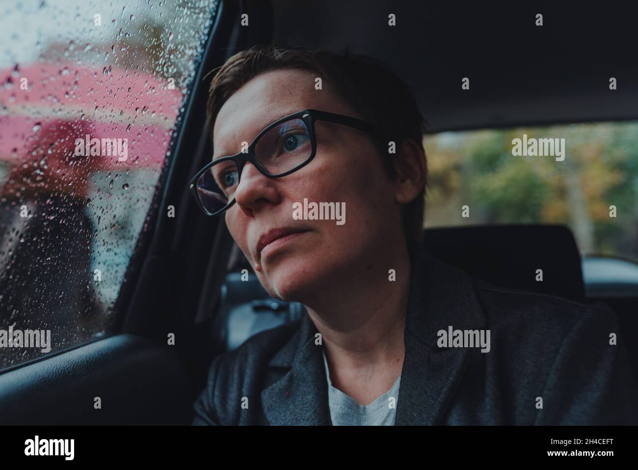 Besorgte Geschäftsfrau mit Brillen, die im Auto warten und bei Regen aus dem Fenster schauen, selektiver Fokus Stockfoto