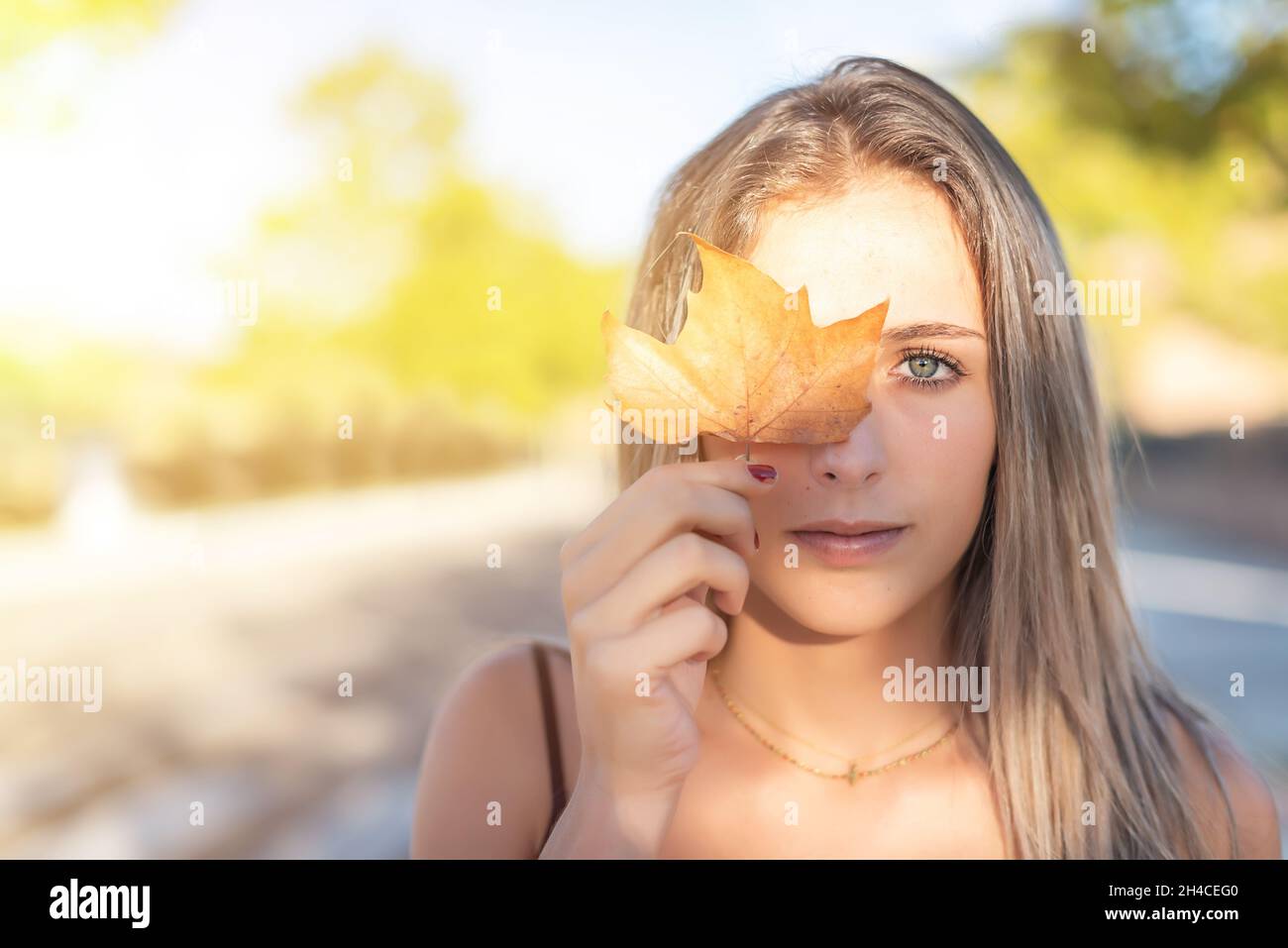 Hübsches junges blondes Mädchen mit grünen Augen, die ein Auge mit einem trockenen Blatt bedecken. Konzept der Ankunft im Herbst Stockfoto