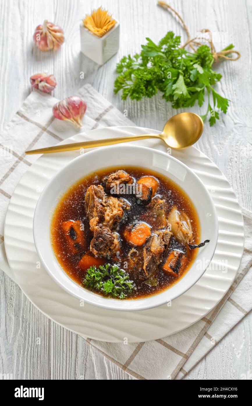 Rinderbrühe von Rinderfleisch auf Knochen langsam mit verkohltem Gemüse gekocht: Karotte, Zwiebel, Knoblauch und Gewürze in einer weißen Schüssel auf einem Holztisch serviert, vert Stockfoto