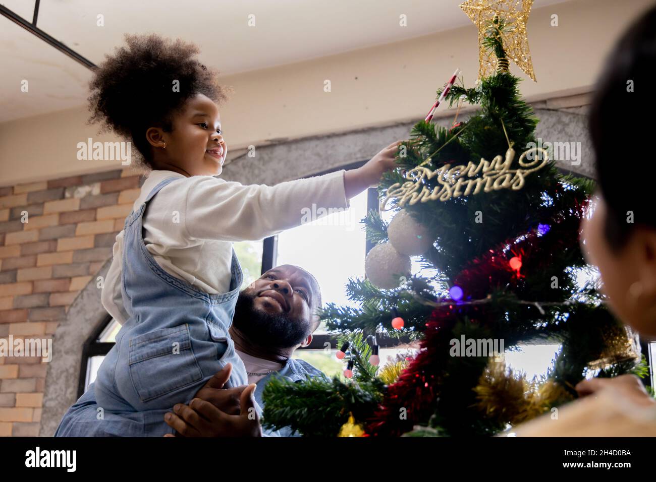 Schönes junges afroamerikanisches Mädchen, das vom hübschen Vater gepflückt wird, der während des Festivals den weihnachtsbaum verschönt, indem er Zuckerrohr an der Spitze des Baumes in der Nähe eines Sterns zu Hause hinzufügt Stockfoto
