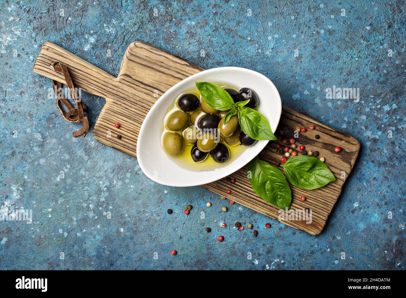 Draufsicht auf leckere Mischung aus grünen und schwarzen Oliven mit Basilikumblättern, Pfeffermischung auf Holzbrett und blauem Beton-Hintergrund Stockfoto