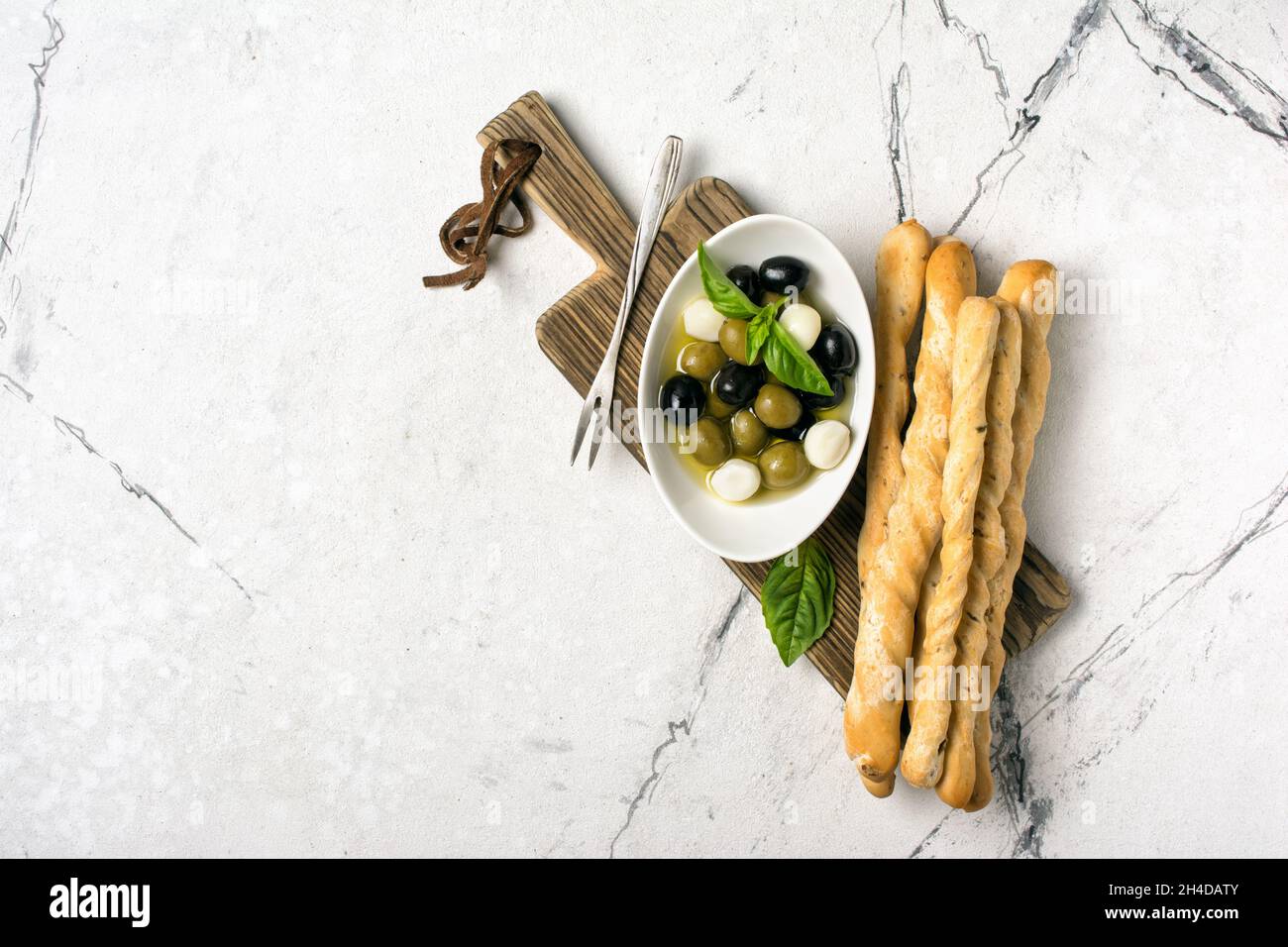 Ansicht von oben auf die Grissini mit Basilikumgewürz, grüne und schwarze Oliven in Öl, Mozzarella auf Holzbrett und weißem Marmorhintergrund mit Kopierfläche Stockfoto