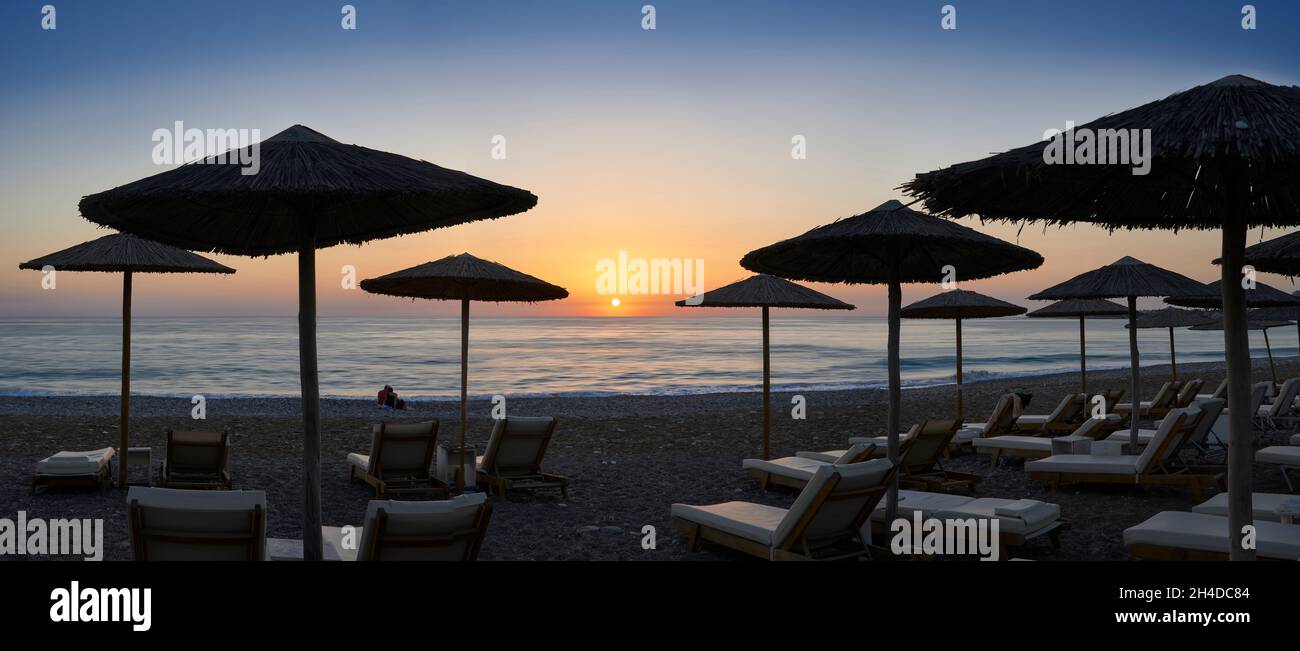 Panoramafoto von Sonnenschirmen und Liegestühlen am Strand in Coral Bay, Zypern bei Sonnenuntergang Stockfoto