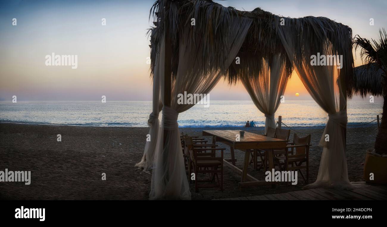Romantischer Tisch unter dem Dach am Strand mit Blick auf das Mittelmeer und Blick auf den Sonnenuntergang Stockfoto