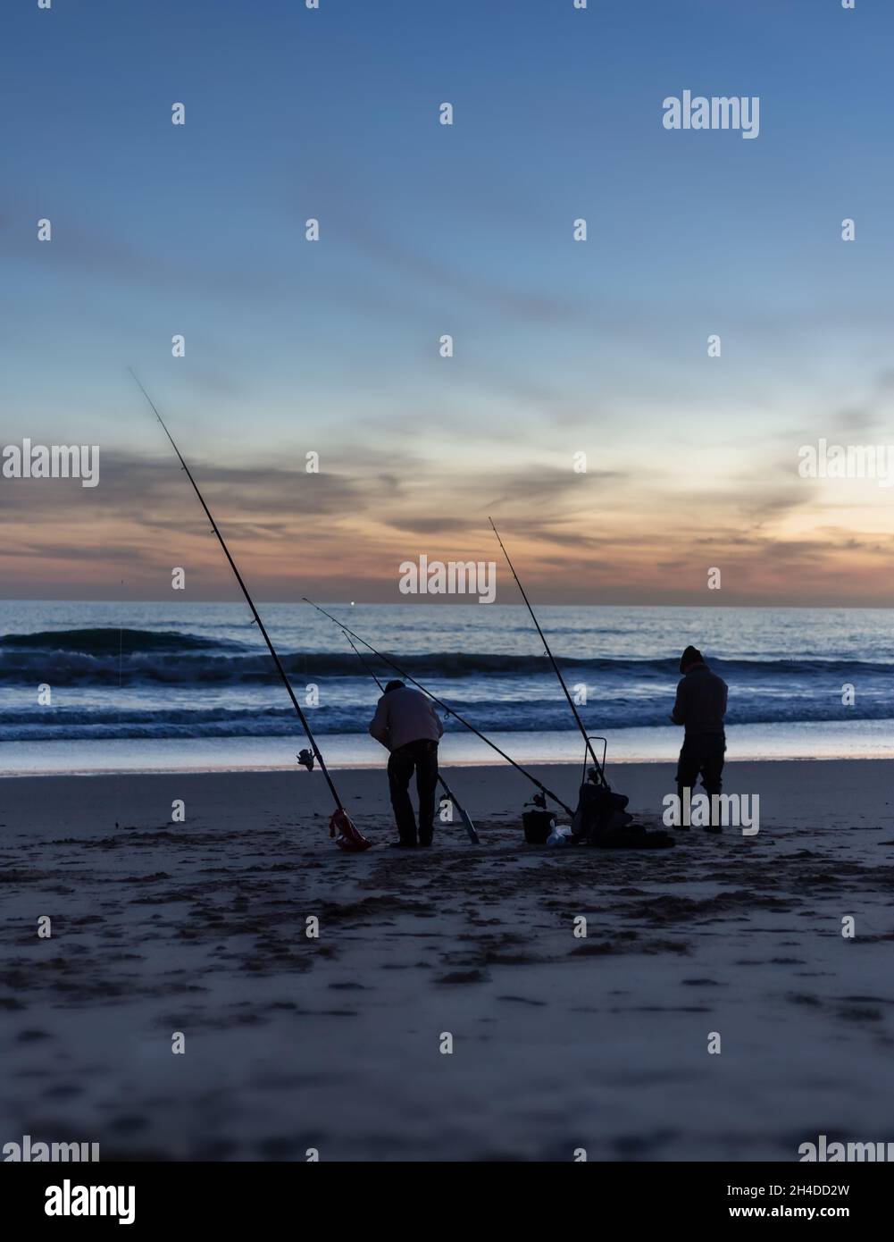 Silhouetten von Fischern mit Spinnruten am Meeresstrand, Angeln im Atlantischen Ozean, am Abend vor dem Hintergrund eines wunderschönen Sonnenuntergangs. Stockfoto