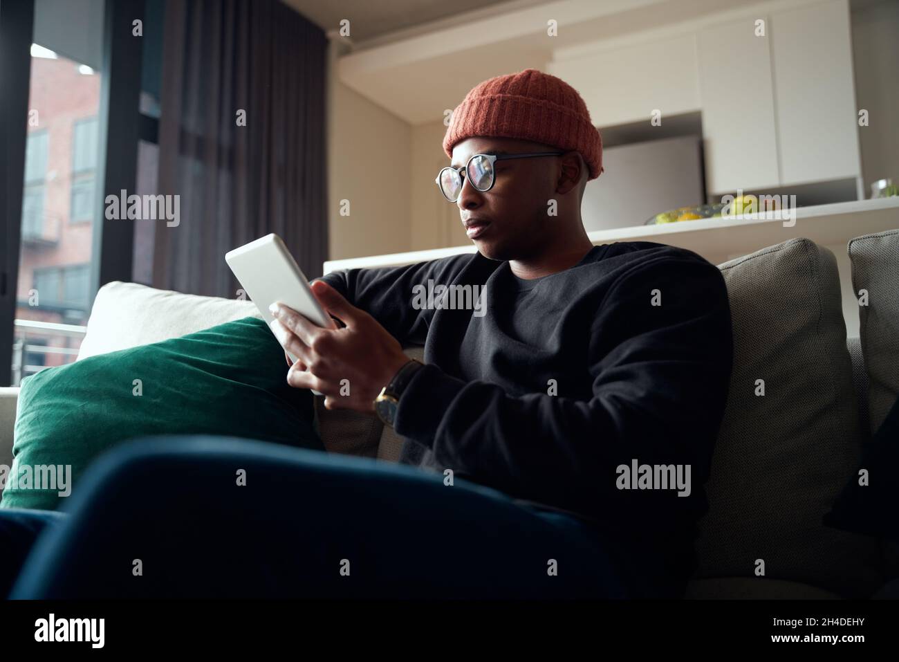 Niedriger Winkel des erwachsenen schwarzen Mannes, der sich beim Betrachen der Tablette konzentriert. Moderne Wohnung auf dem Sofa Stockfoto