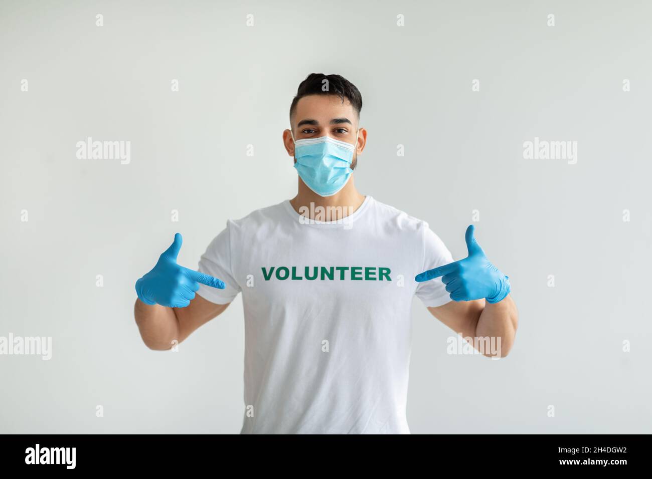 Sehr stolz, ehrenamtlich zu sein. Junger arabischer Mann mit medizinischer Maske und Handschuhen, die auf seine Uniform zeigen, über einer hellen Wand positioniert Stockfoto