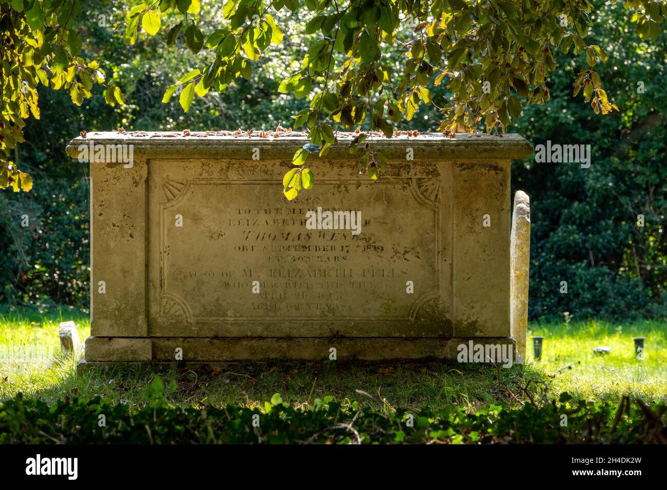 Ein Grab im Gewölbestil, das im Schatten eines Baumes mit einem hellen Sonneneinstrahlung hinter der St. Lawrence Church Knodishall, Suffolk, zu sehen ist Stockfoto