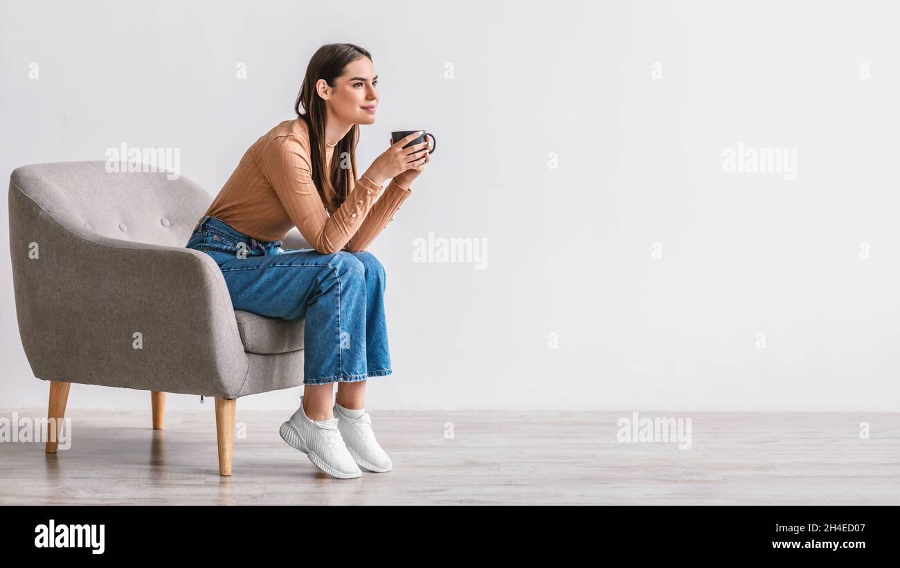 Verträumte junge kaukasische Frau trinkt Kaffee, Pause im Sessel gegen weiße Studiowand, Banner mit Kopierraum Stockfoto