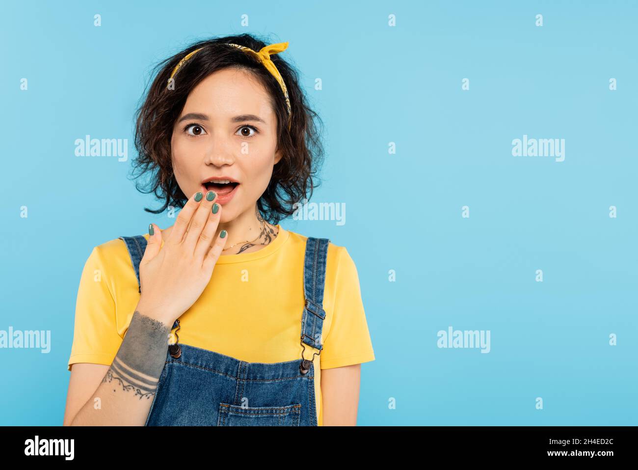 Staunend tätowierte Frau, die offenen Mund mit Hand isoliert auf blau bedeckt Stockfoto