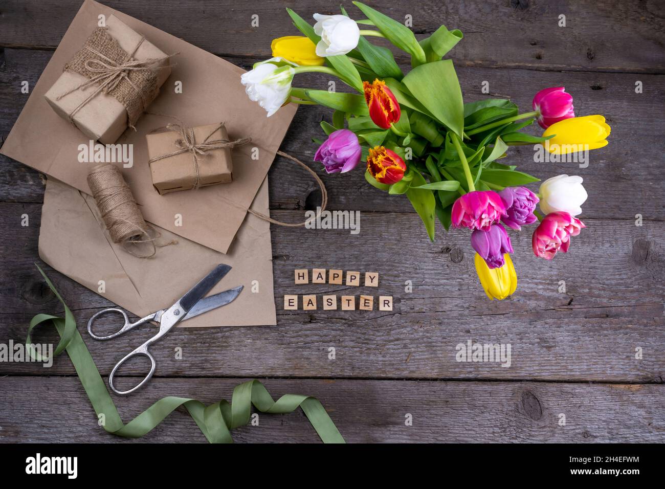Frohe Ostereinschrift in Holzbuchstaben. Geschenkverpackung. Mix aus Frühlingsblumen. Hintergrund mit Blumen Tulpen close-up verschiedenen Farben. Geschenk. Draufsicht. Speicherplatz kopieren. Handgemacht Papper. Stockfoto