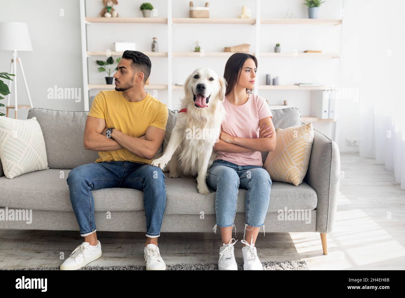 Unglücklich beleidigte das junge, vielfältige Paar, das in entgegengesetzte Richtungen schaute und auf dem Sofa saß, mit einem niedlichen Hund dazwischen Stockfoto
