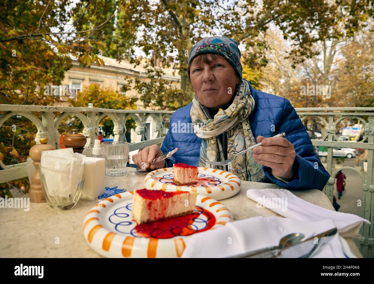 Schöne reife Erwachsene Frau, die Käskuchen an einem Restauranttisch auf dem Balkon mit Herbstbäumen im Hintergrund isst. Stockfoto