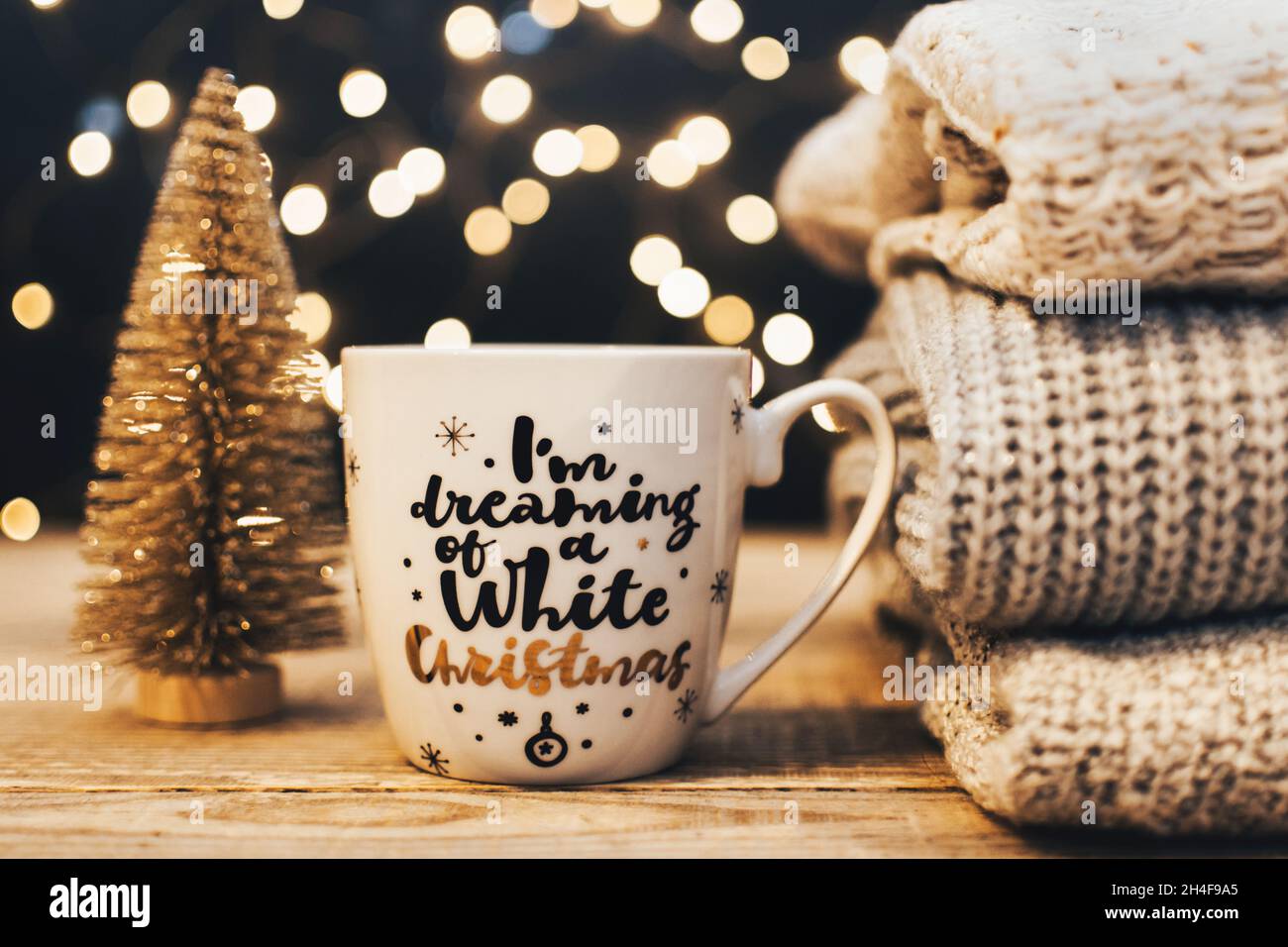 Weihnachtsstill-Life-Bild mit Stapel von warmen Winterpullover, weißem weihnachtsbecher mit Text und dekorativem weihnachtsbaum vor Bokeh-Lichtern Stockfoto