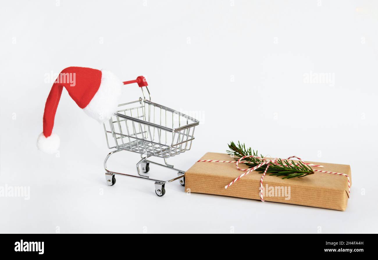 Mini-Einkaufswagen mit weihnachtsmütze und eingewickeltem weihnachtsgeschenk auf weißem Hintergrund mit Kopierfläche Stockfoto