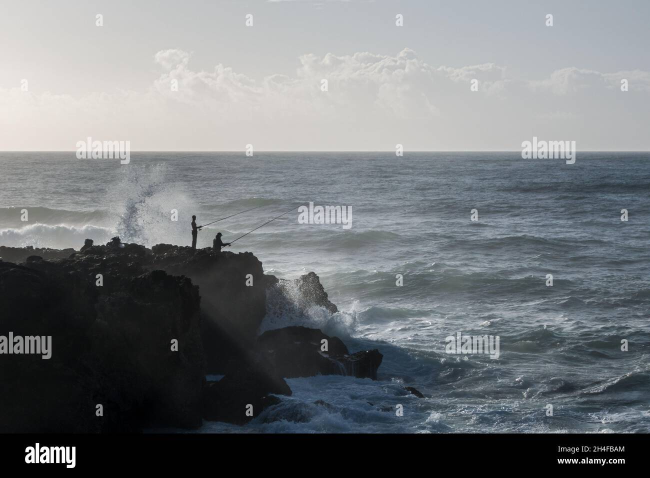 Zwei Sportfischer mit Stangen, die auf einem Felsvorsprung am Atlantischen Ozean stehen, mit rauer See und Wellen, die sich einrollen, wobei sich das Spray an der Küste aufspüllt Stockfoto