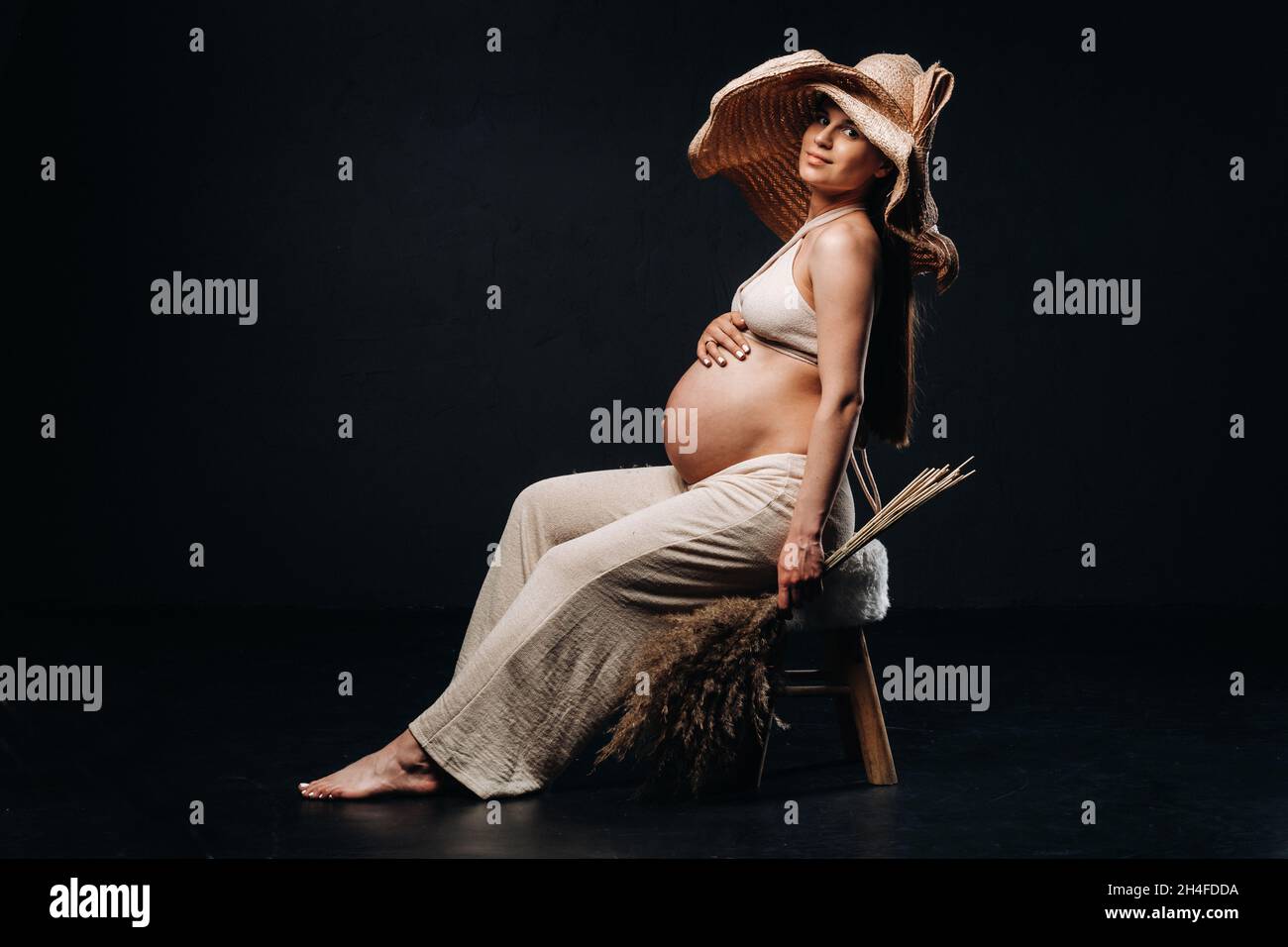 Eine Schwangere in einem Strohhut mit beigen Kleidern und einem Strauß in den Händen im Studio auf schwarzem Hintergrund. Stockfoto