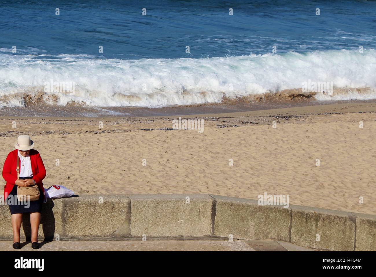 Eine Dame (keine Erkennbarkeit) mit einem roten Blazer, schwarzem Rock und weißem Hut sitzt mit einer Handtasche auf dem Schoß auf einer Strandmauer. Stockfoto