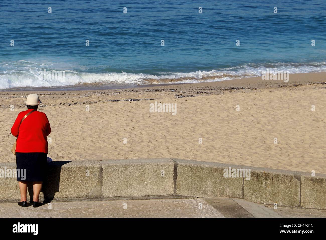 Eine ältere Dame (keine Erkennbarkeit) mit einem roten Blazer, einem schwarzen Rock und weißem Hut steht vor einer Strandmauer, Espinho, Portugal. Stockfoto