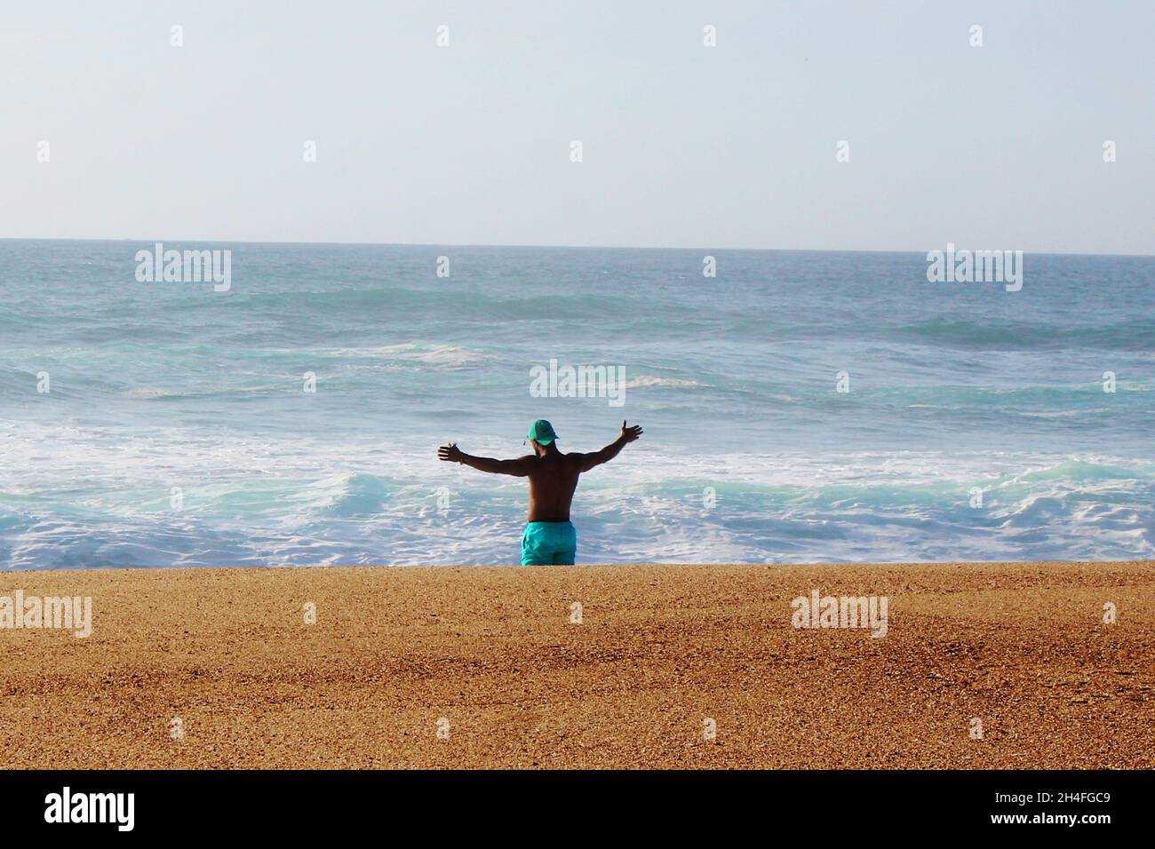 Schwarzer Mann (keine Erkennbarkeit), der eine Shorts und ein Cap in mint tragt, breit seine Arme aus, über den Wellen und am Strand von Nazare. Stockfoto