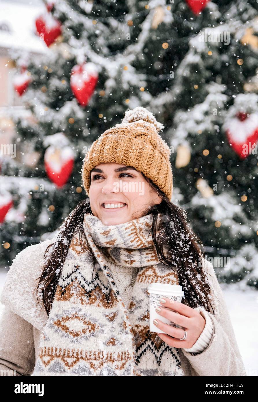 Porträt einer jungen Frau, die an einem verschneiten Tag in der Stadt stilvolle Winterkleidung trägt und unterwegs Kaffee trinkt Stockfoto