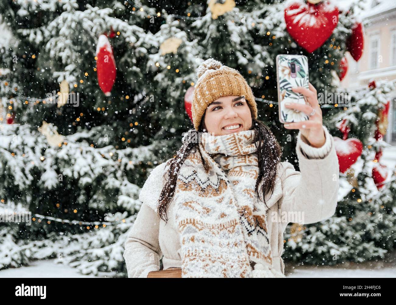 Porträt einer schönen, glücklichen jungen Frau, die am verschneiten Tag in der Stadt ein Selfie vor dem weihnachtsbaum macht Stockfoto