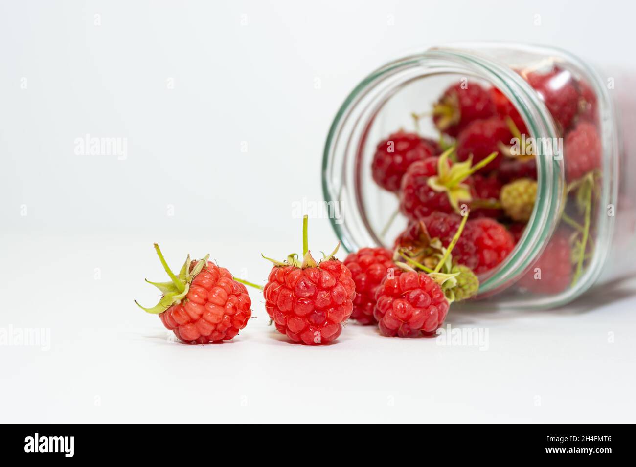 Frisch gepflückte rote Himbeeren, einige Beeren aus einem Glas auf weißem Grund verstreut. Stockfoto