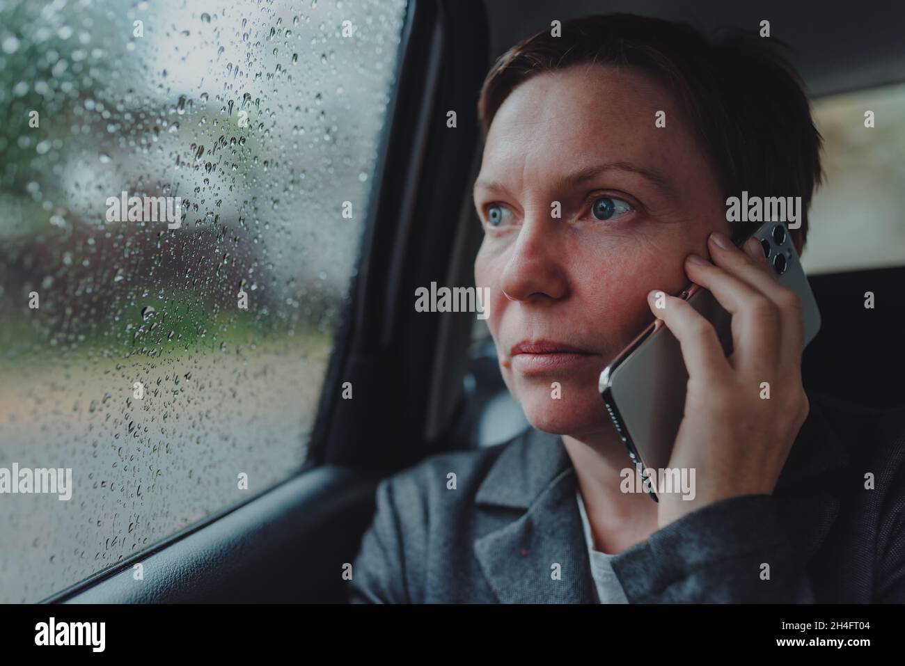 Seriöse Geschäftsfrau, die während des Regens auf dem Rücksitz des Autos über das Mobiltelefon spricht, selektiver Fokus Stockfoto