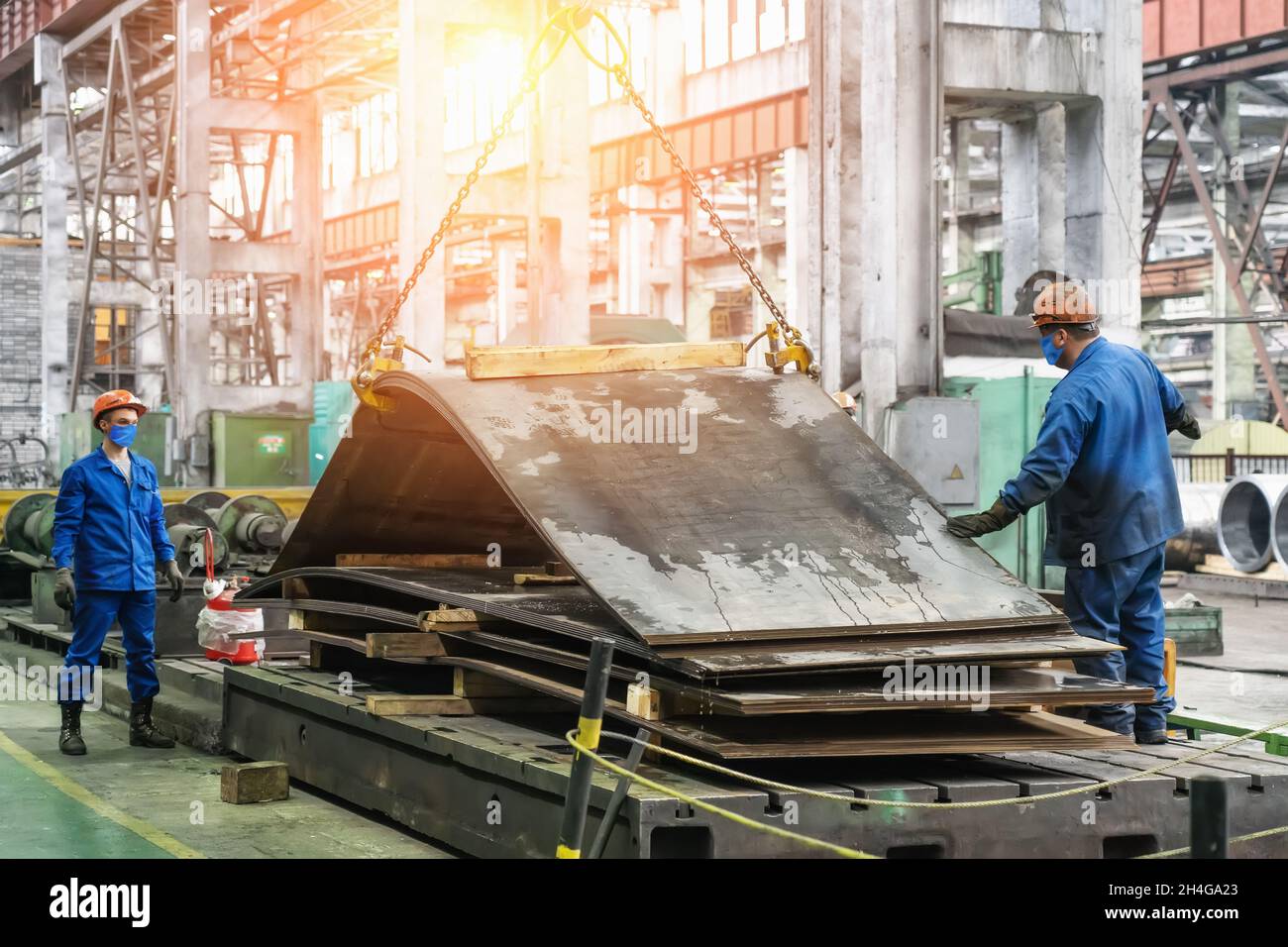 Arbeiter in Masken und Helmen im metallurgischen Werk entladen Stahl- oder Metallbleche mit einem Kran für die Stahlproduktion. Stockfoto