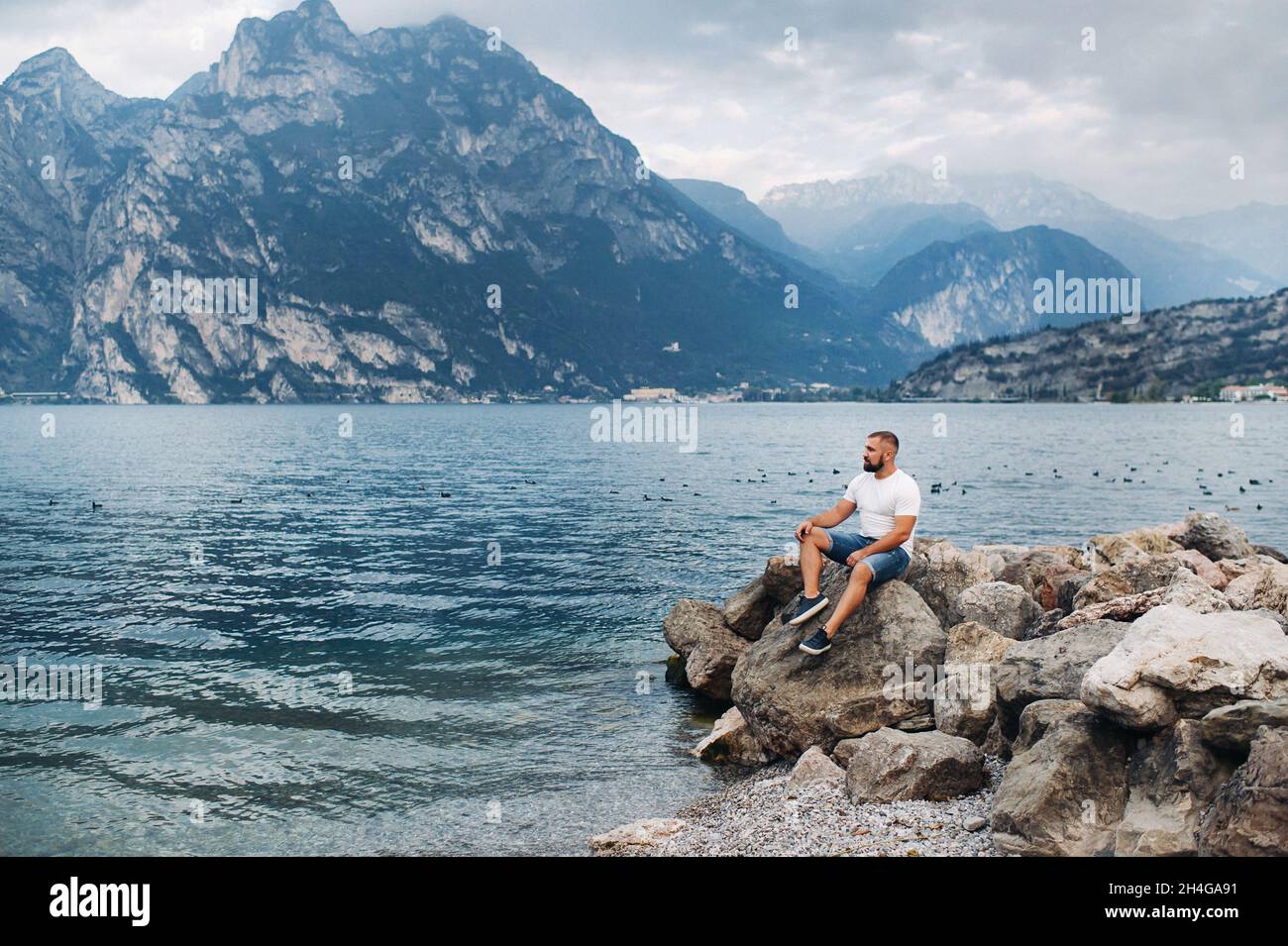 Männlicher Reisender sitzt auf den Felsen vor der Kulisse der Alpen und des Gardasees. Urlaub in Italien. Stockfoto