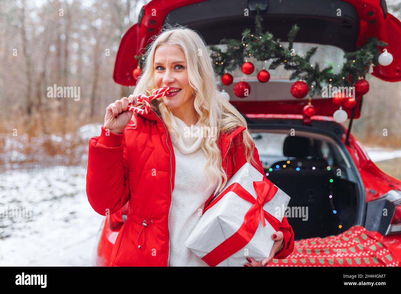 Lächelnde blonde Frau in roter Jacke mit herzförmigen roten und weißen Bonbons halten Geschenkbox mit rotem Band in der Nähe weihnachten dekorierten Auto im Winter Stockfoto