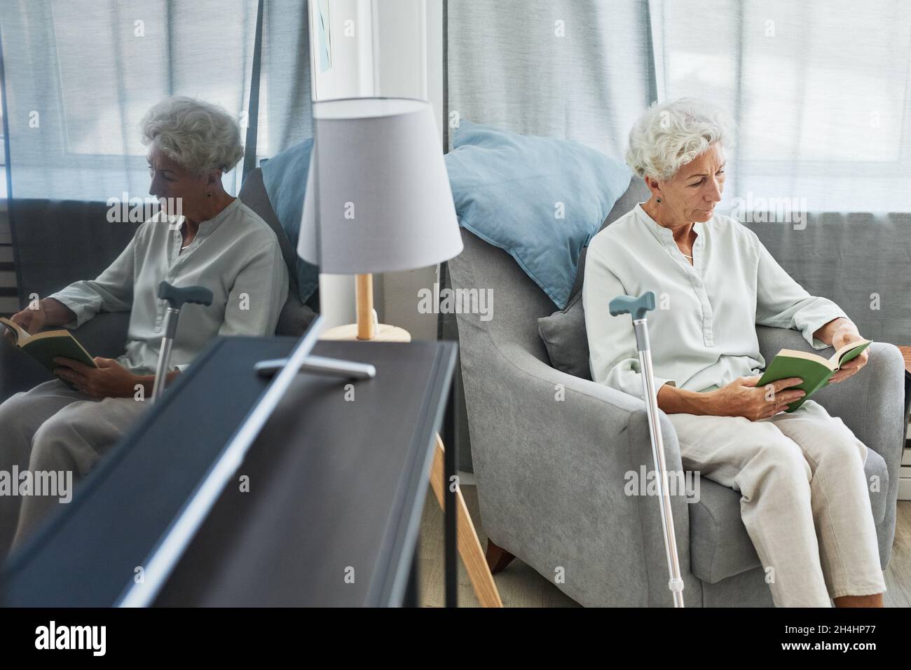 Hochwinkelporträt einer weißhaarigen älteren Frau, die zu Hause Buch liest, während sie im Sessel sitzt, mit Spiegelung aufgenommen, Platz zum Kopieren Stockfoto