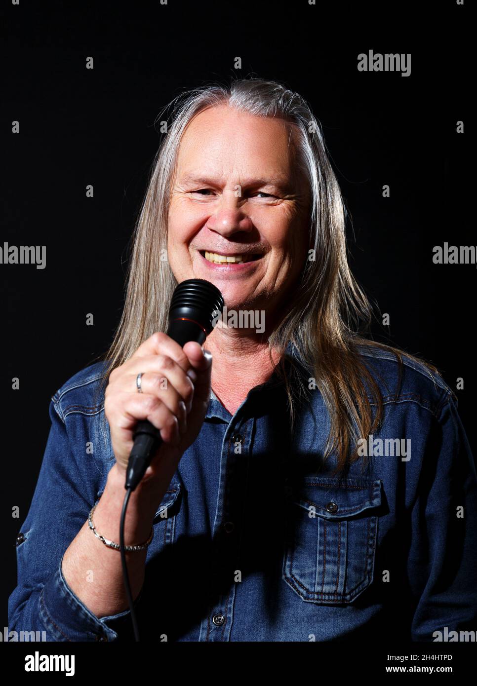 Älterer Mann mit langen grauen Haaren in blauem Denim-Hemd singt auf schwarzem Hintergrund in ein Mikrofon auf der Bühne Stockfoto