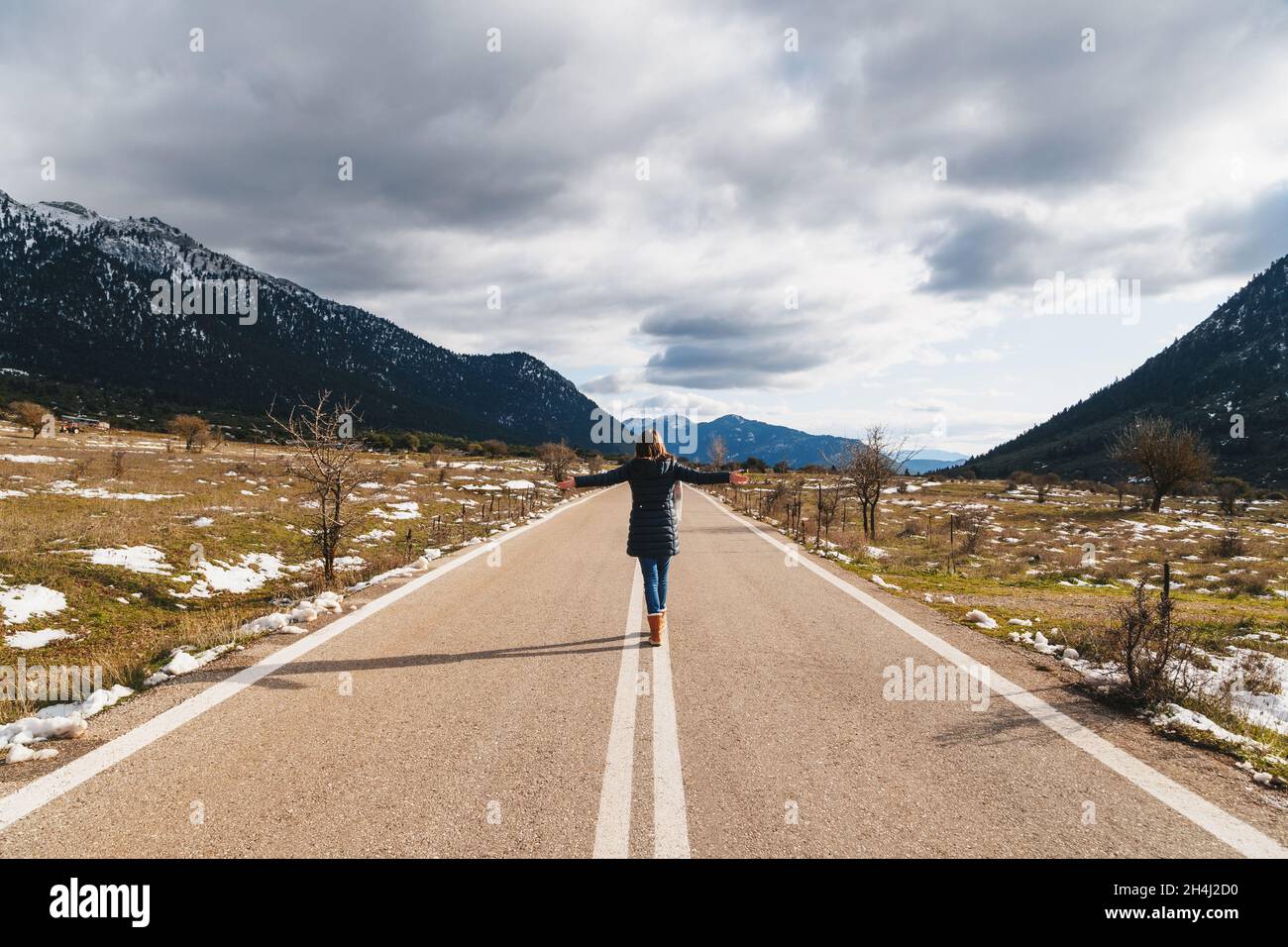 Rückansicht einer jungen Frau, die mit erhobenen Händen auf einer leeren Asphaltstraße mitten in einer bergigen Gegend läuft. Hochland von Griechenland, Winterzeit. Freiheit, Reisekonzept. Stockfoto