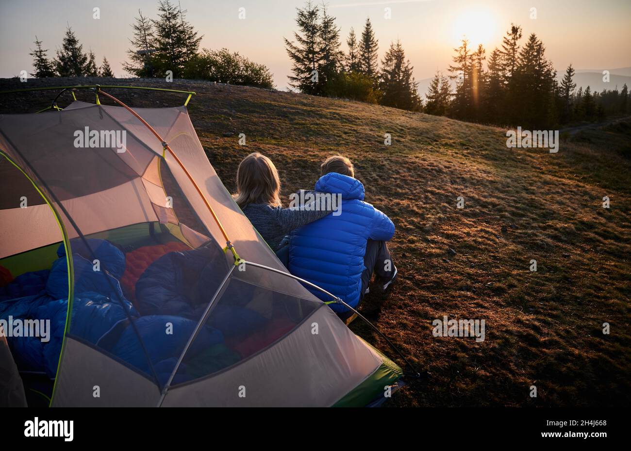 Rückansicht von zwei Reisenden, Mann und Frau, die auf Gras vor dem Zelt auf einem Berg sitzen, früh am Morgen und über den sonnigen Himmel nachsinnen. Konzept von Wandern, Camping und Beziehungen. Stockfoto