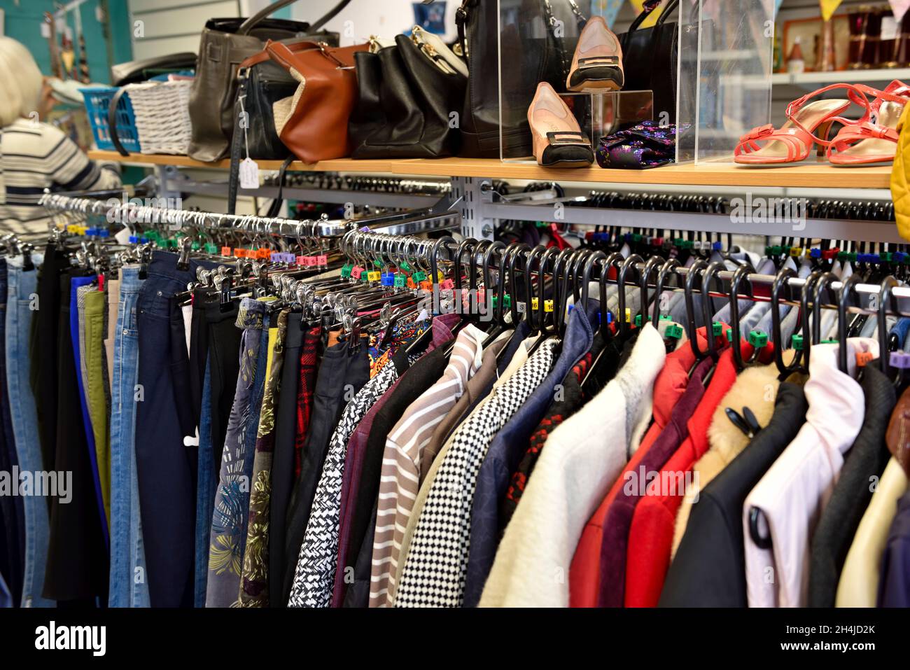 Gebrauchte Tücher zum Verkauf, die an der Schiene mit Taschen und Schuhen im Wohltätigkeitsgeschäft, Henley-in-Arden, Großbritannien, hängen Stockfoto