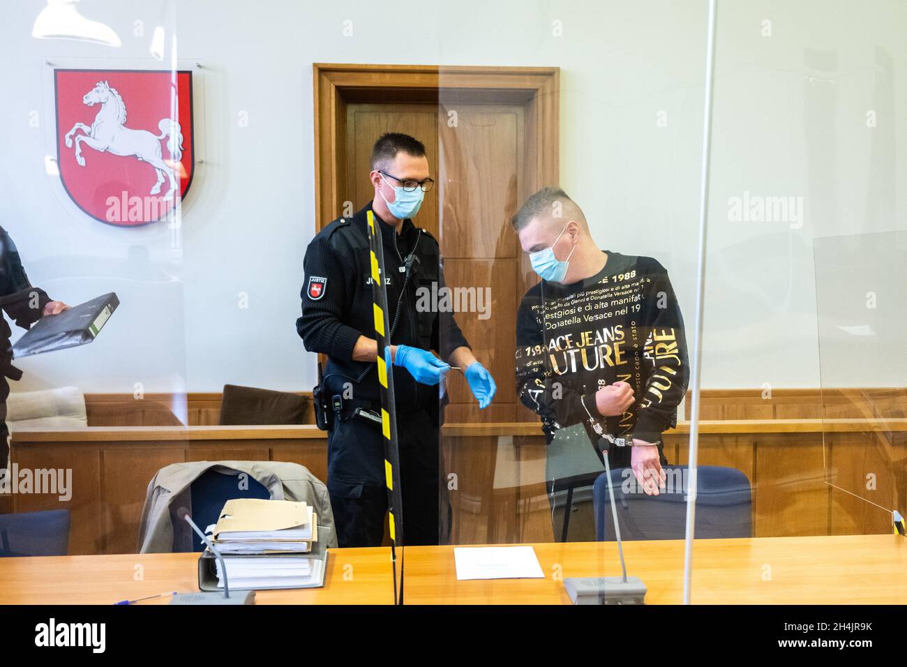 Oldenburg, Deutschland. November 2021. Der Angeklagte (r) steht in Handschellen neben einem Gerichtsbeamten, während die Verhandlung im Gerichtssaal des Landgerichts beginnt. Er soll gefälschte Goldbarren und Krügerrand-Münzen über eine Online-Plattform verkauft haben. Dabei soll er Schäden von rund 40,000 Euro verursacht haben. Kredit: Mohssen Assanimoghaddam/dpa/Alamy Live Nachrichten Stockfoto