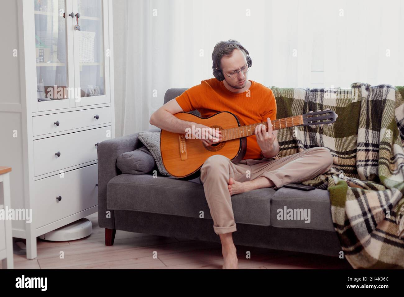 Junger Kaukasischer mit Kopfhörern, der Gitarre spielt und zu Hause auf der Couch sitzt. Üben Sie Musik, Lifestyle-Foto. Stockfoto