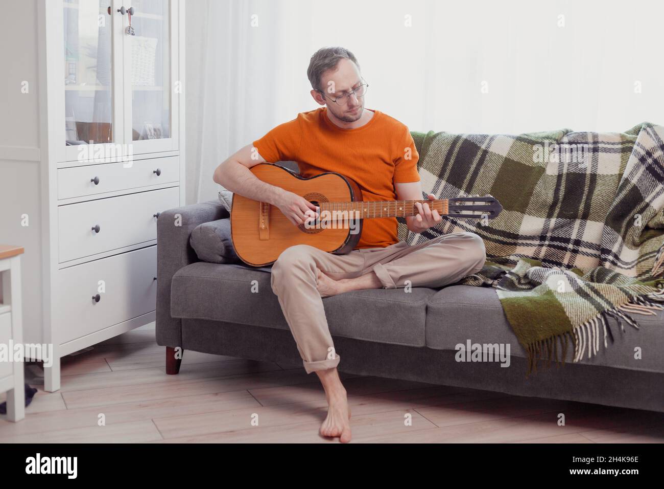 Der junge Kaukasusmann spielt Gitarre und sitzt zu Hause auf der Couch. Üben Sie Musik, Lifestyle-Foto. Stockfoto