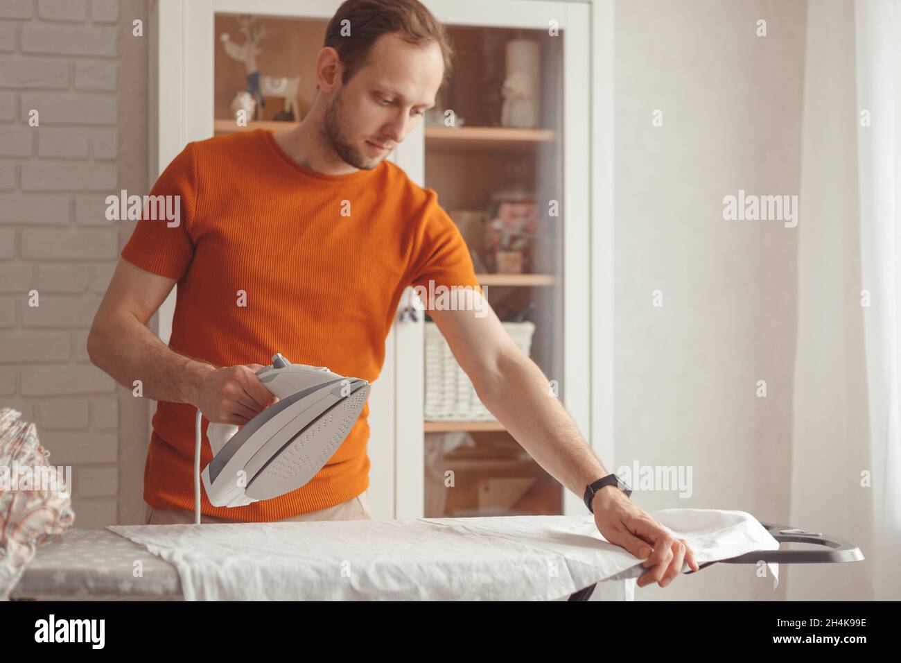 Der junge Kaukasusmann bügelt zu Hause das Kindertuch auf dem Bügelbrett. Männer, die Hausarbeiten erledigen. Stockfoto