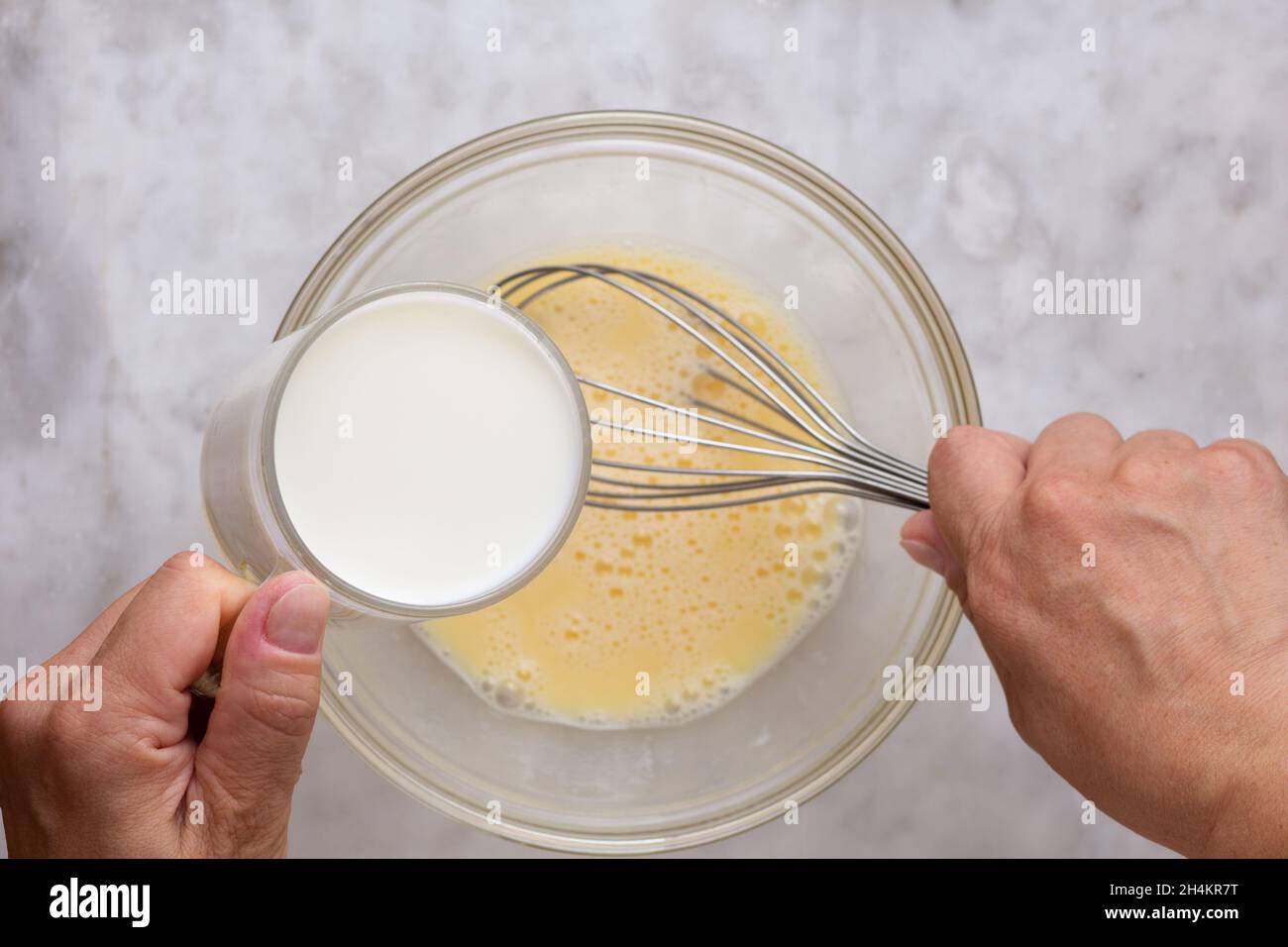 Draufsicht auf eine Frau, die ein Glas Milch in einer Glasschüssel auf der Marmoroberfläche in gemischte rohe Eier legt Stockfoto