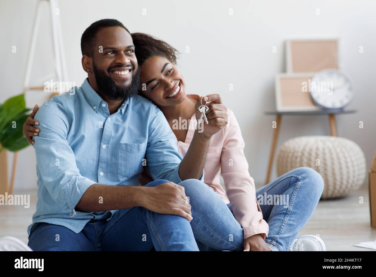 Froher, tausendjähriger afroamerikanischer Ehemann und Ehefrau zieht in eine neue Wohnung, sitzt zwischen Kisten und zeigt Hausschlüssel in einem neuen Zuhause. Versicherung, Immobilien, m Stockfoto