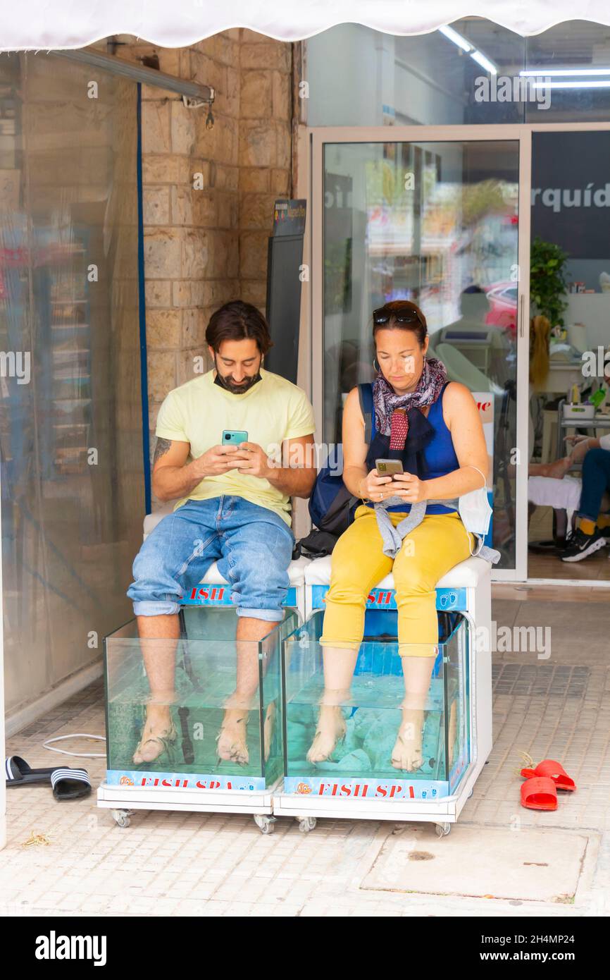 Das Paar sitzt auf dem Telefon und lässt sich in Can Pastilla Mallorca Spanien von Fischen die Füße knabberen Stockfoto