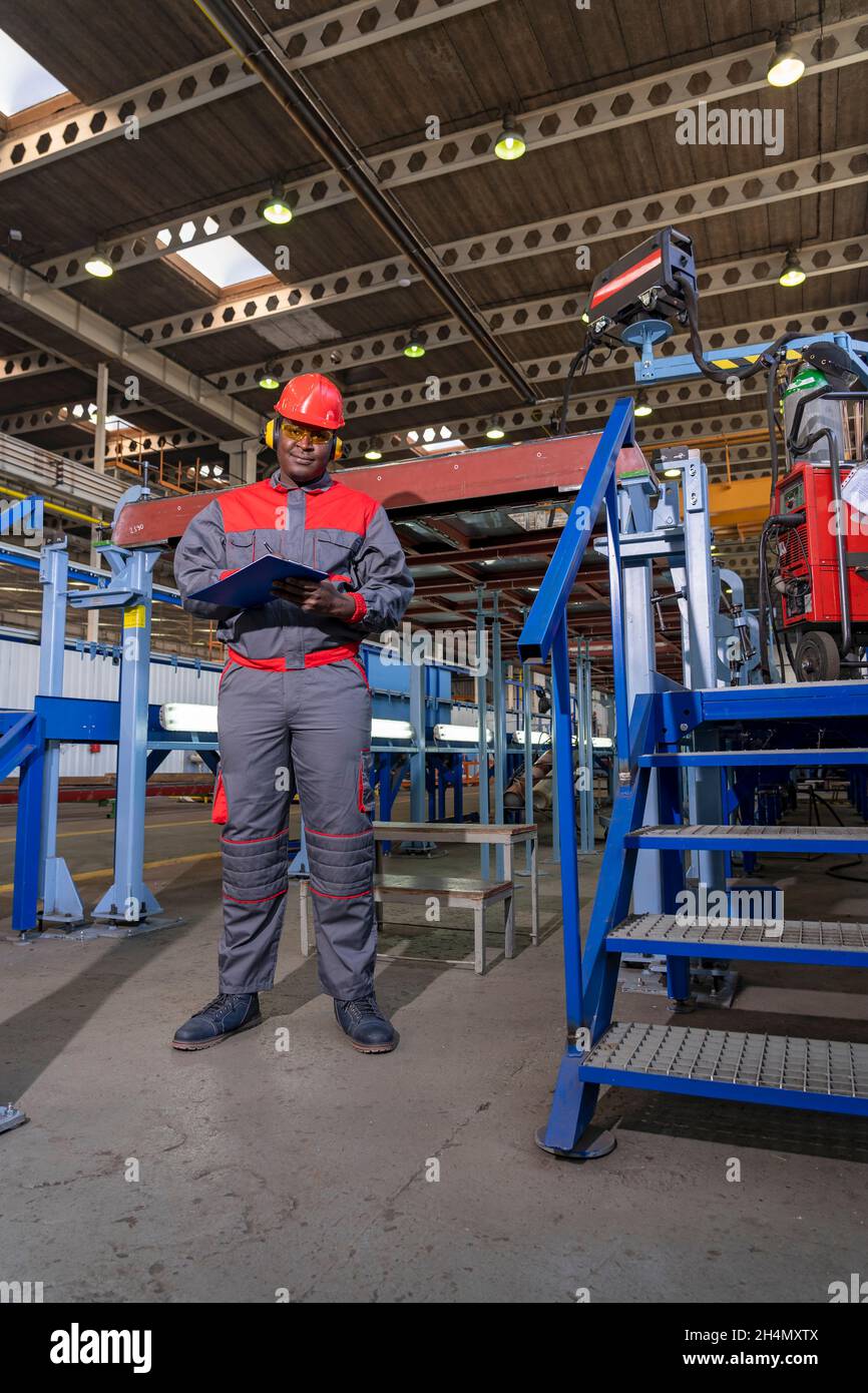 Full Figure Portrait of Black Industrial Worker in Red Helmet, Yellow Safety Goggles and work uniform in A Factory. Stockfoto