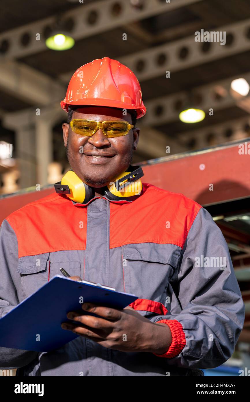 Waist Up Portrait von schwarzen Industriearbeitern in rotem Helm, gelben Schutzbrillen und Arbeitskleidung in Einer Fabrik. Stockfoto