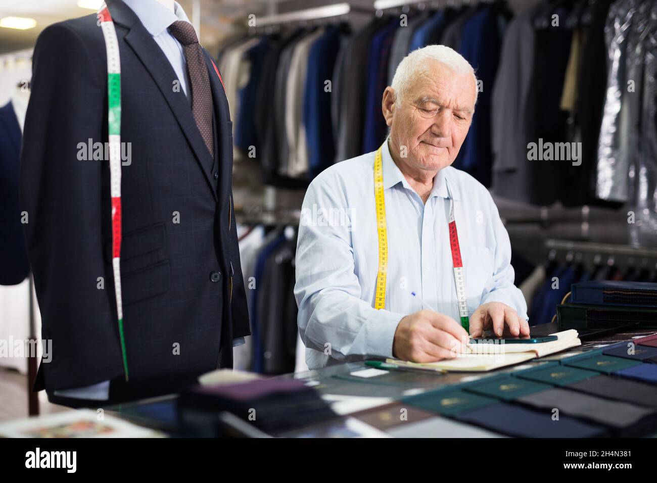 Der ältere Schneidermann nimmt Messungen aus seiner Jacke und schreibt sie in der Werkstatt in ein Notizbuch Stockfoto
