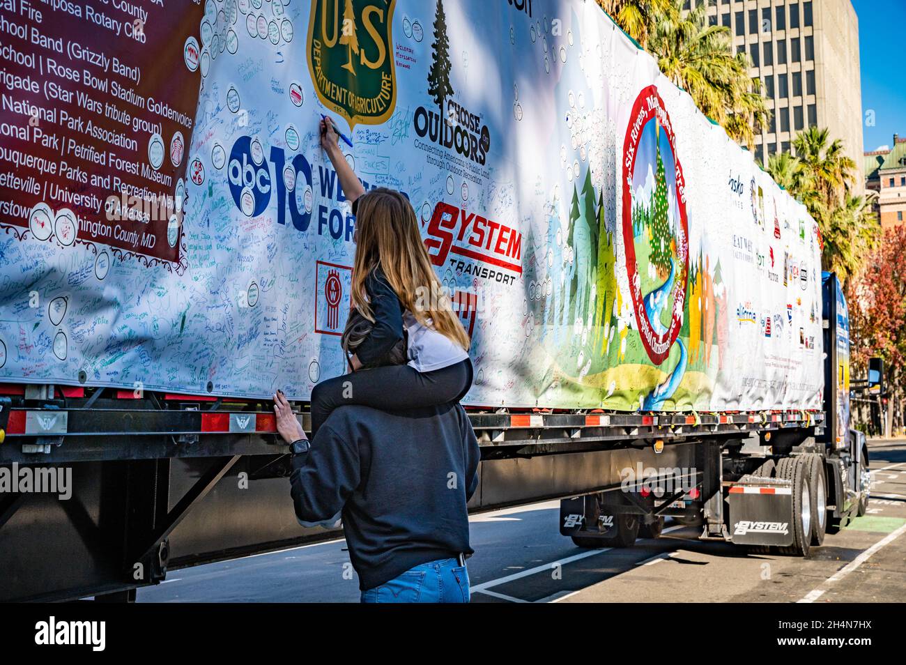 Ein Mann hält ein Mädchen auf seinen Schultern, während sie auf dem Schild auf der Seite des Kenworth T680-Trucks schreibt, der den Weihnachtsbaum des US-Kapitols nach D.C. bringt Stockfoto