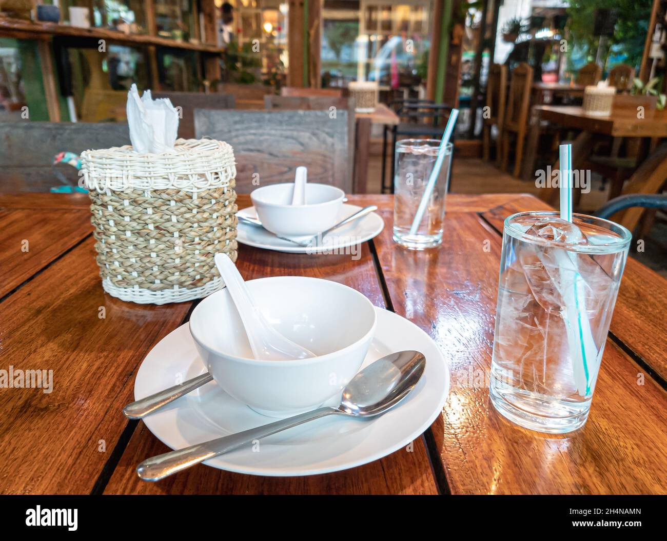 Thai-Restaurant mit Tischeinrichtung. Gläser Wasser mit Stroh, Tellern und Seidenpapier auf Holztisch. Stockfoto